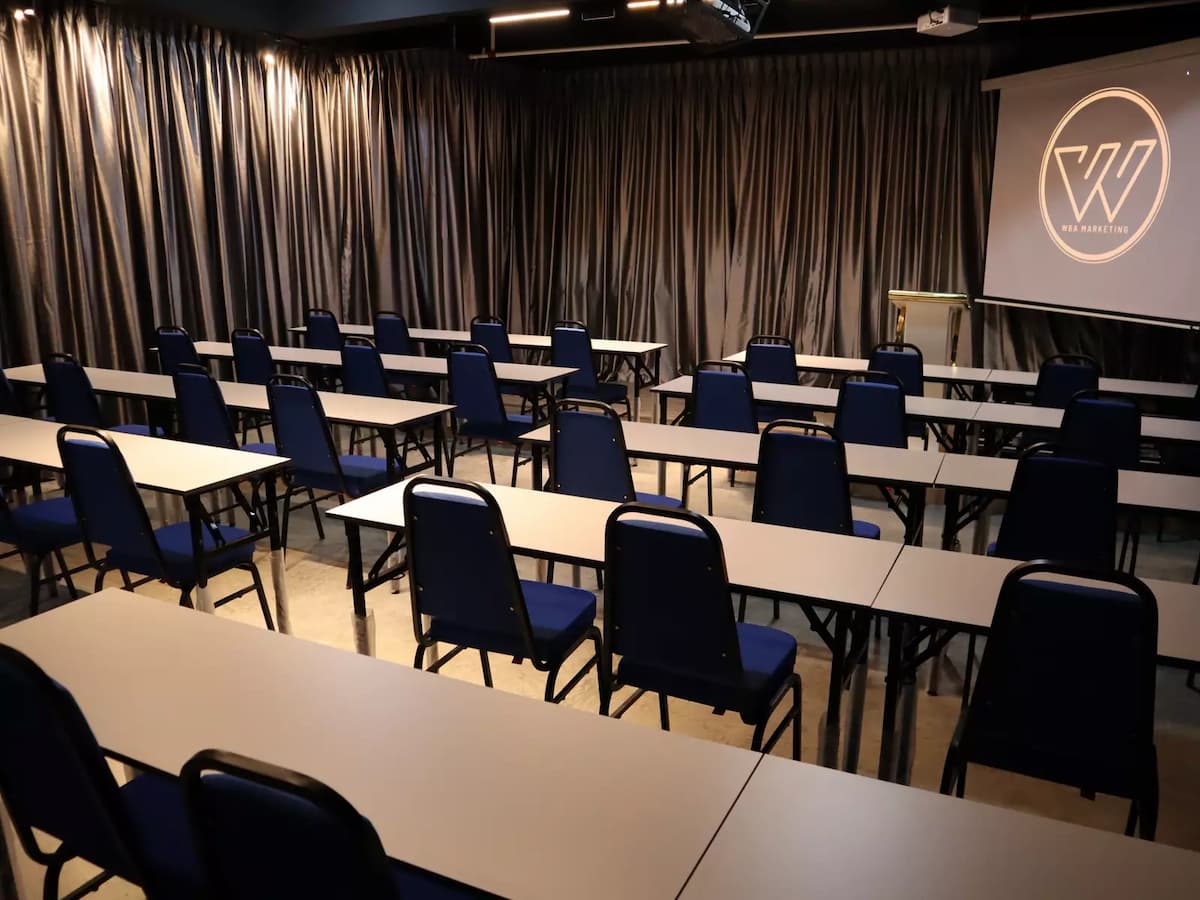 rows of white tables and blue chairs facing a projector screen and podium