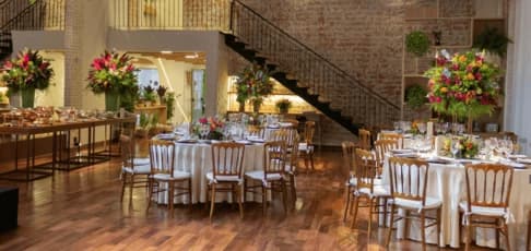 Banquet-style event space with round dining tables, wooden chairs, tall floral centerpieces, polished wooden floors, and exposed brick walls, set up for a formal dinner or private event inside a multi-level venue.