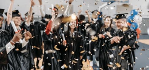Group of graduates in caps and gowns cheering and tossing confetti during a graduation party celebration.