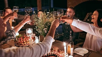 A close-up view from above the table of a diverse group of friends raising their wine glasses in a toast at a candlelit outdoor dinner table at night. The table is set with a large bouquet of wildflowers, appetizer skewers, and wine bottles.