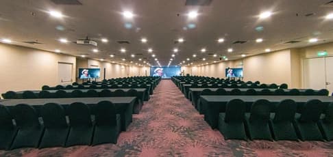 Wide-angle view of the large, empty conference hall at PEN & INC arranged in a classroom style. Symmetric rows of long black tables and chairs are separated by a central carpeted aisle, featuring a large LED stage screen and side monitors.