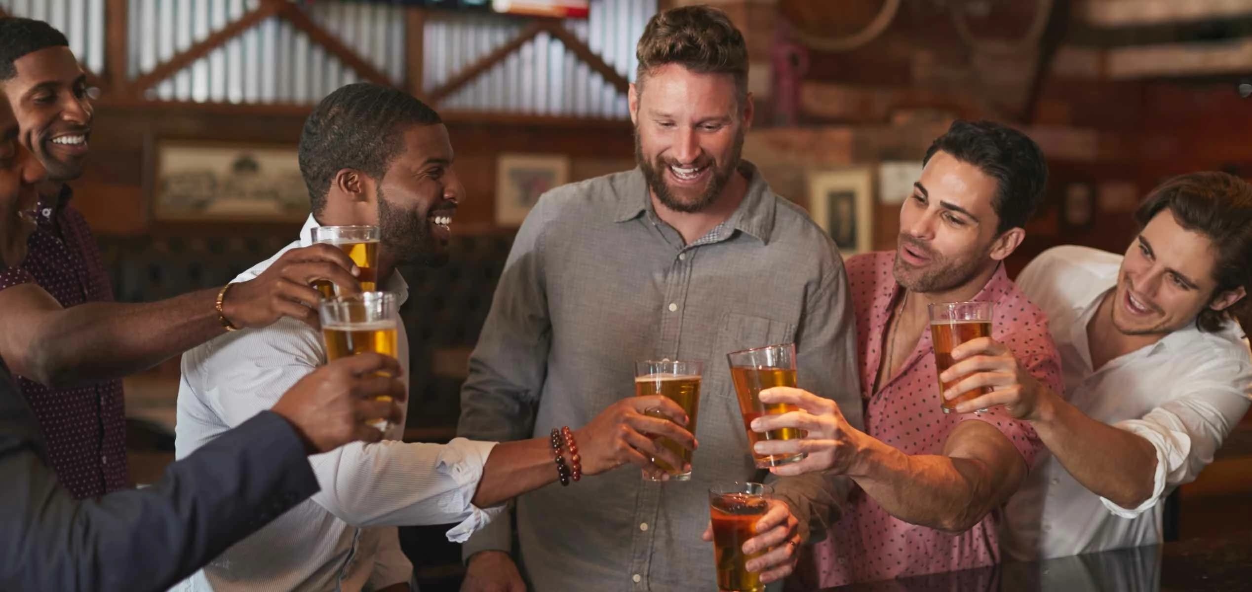A diverse group of six smiling men laughing and clinking pint glasses filled with amber beer together during a celebratory toast inside a rustic bar environment.