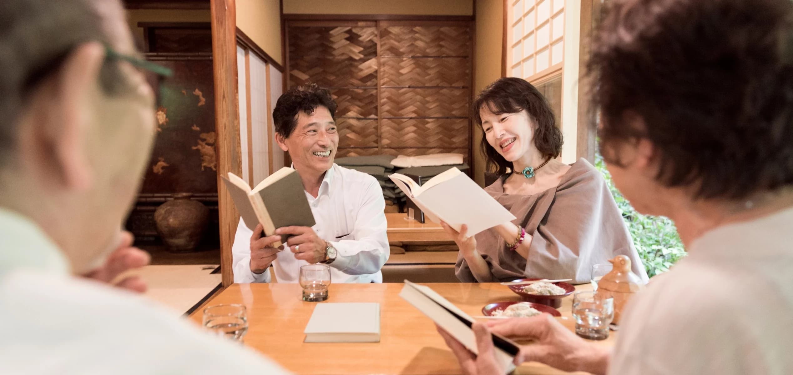 A group of adults smiling and discussing books around a table, representing a lively book club and the power of social community gatherings.