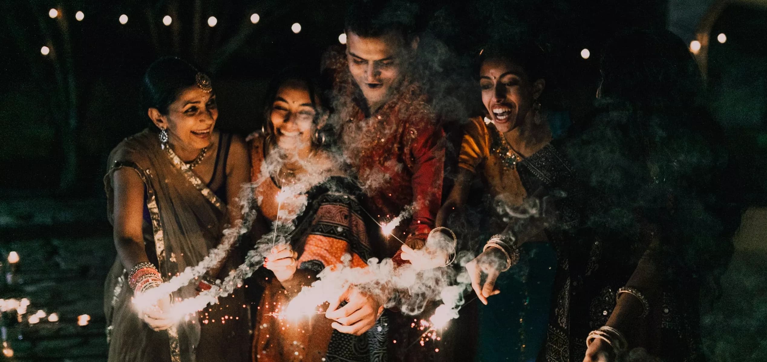 A group of friends joyfully celebrating Diwali outdoors at night. They are dressed in colorful traditional Indian attire and are smiling brightly as they hold lit sparklers, surrounded by trails of festive smoke and illuminated by warm string lights in the dark background.