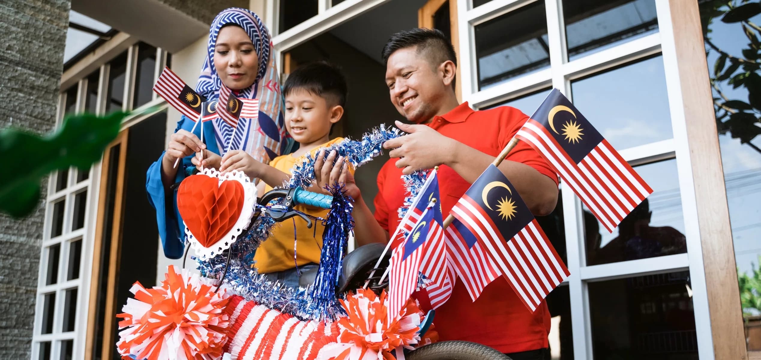 A smiling family of three, including a mother wearing a patterned hijab, a father in a red polo, and their young son, stand outside their home decorating a child's bicycle. They are adorning the bike with small Malaysian flags, shiny blue tinsel, and a red paper heart to celebrate Merdeka.