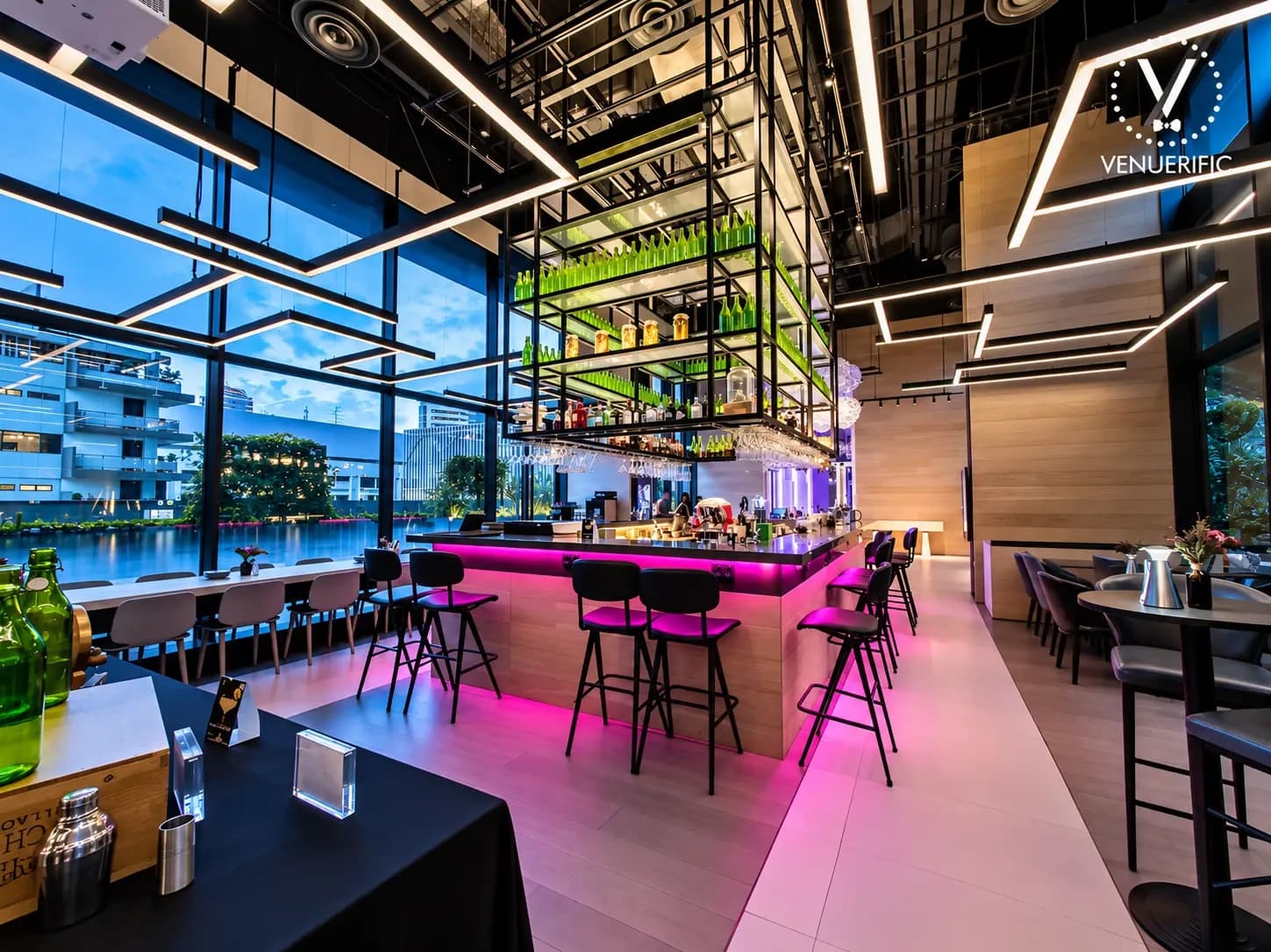 Komyuniti bar with a suspended rack of green bottles, pink-lit counter, black stools and floor-to-ceiling windows at dusk