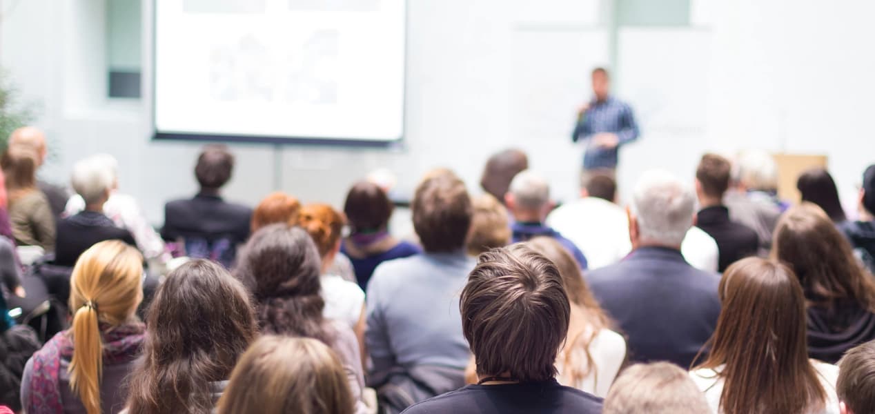 Rows of staff facing a speaker holding a microphone beside a projector screen at a corporate town hall event