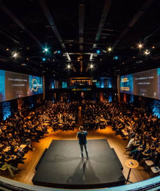 wide view of speaker presenting on center stage in large auditorium with dual projection screens and full audience