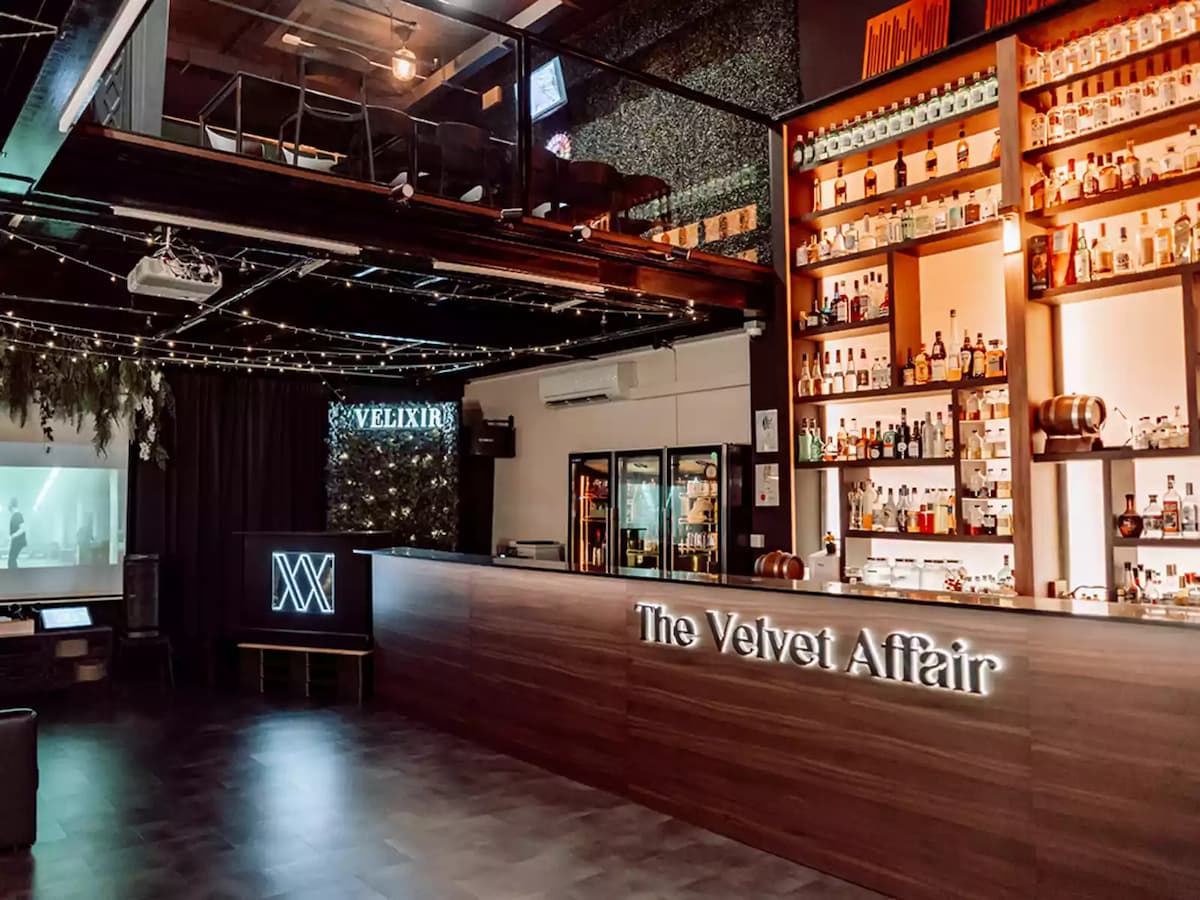 The Velvet Affair venue interior showing the main bar with illuminated spirit shelves, a Velixir branded DJ booth, string lights, projection screen, and mezzanine level above