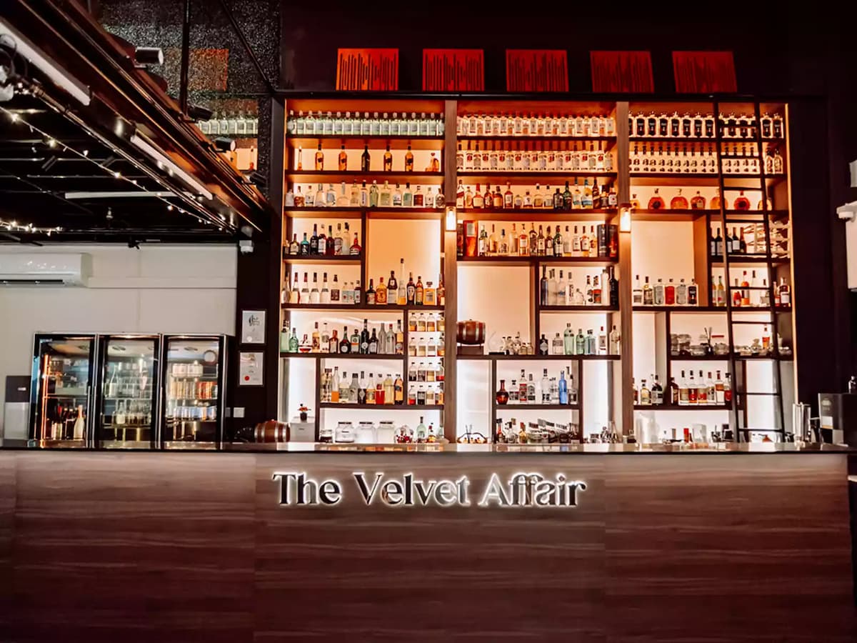 The Velvet Affair bar front with illuminated shelves lined with spirits and liquor bottles, beverage fridges, and the venue name displayed on the bar counter