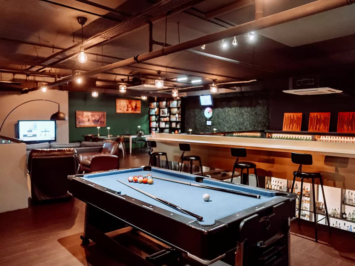 Upscale games bar with a blue felt pool table in the foreground, a long bar counter with bar stools, lounge seating area, and warm Edison bulb lighting throughout