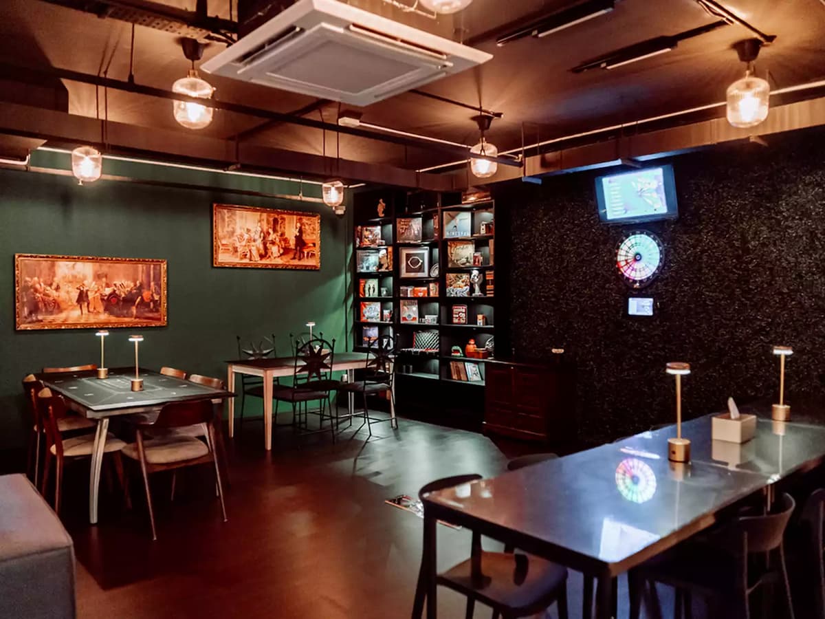 Intimate games lounge with dining tables, a board game bookshelf, electronic dartboard, wall-mounted TV, framed artwork, and warm pendant lighting against dark green walls