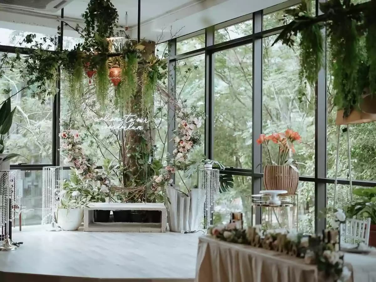 Indoor wedding stage with floral arch, hanging greenery, and floor-to-ceiling windows overlooking trees.