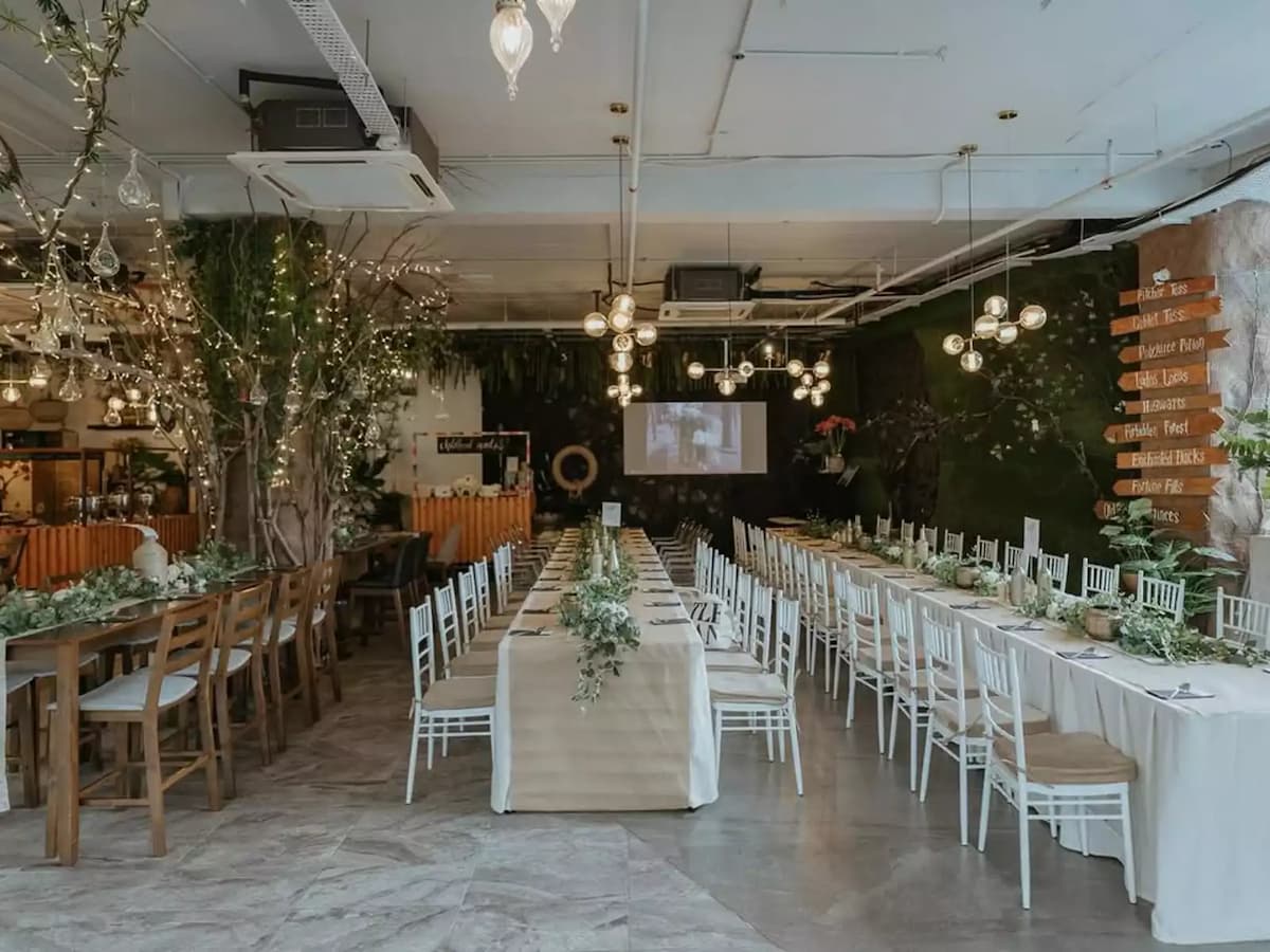 Rustic indoor banquet setup with long tables, greenery decor, string lights, and a presentation screen at the front.