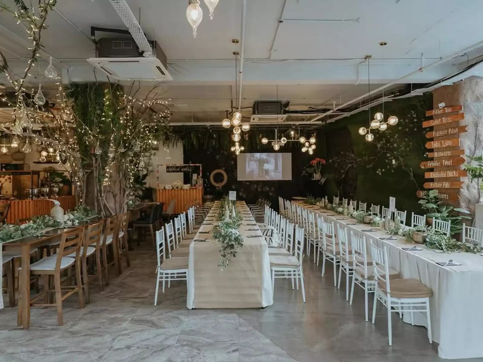 Rustic indoor banquet setup with long tables, greenery decor, string lights, and a presentation screen at the front.