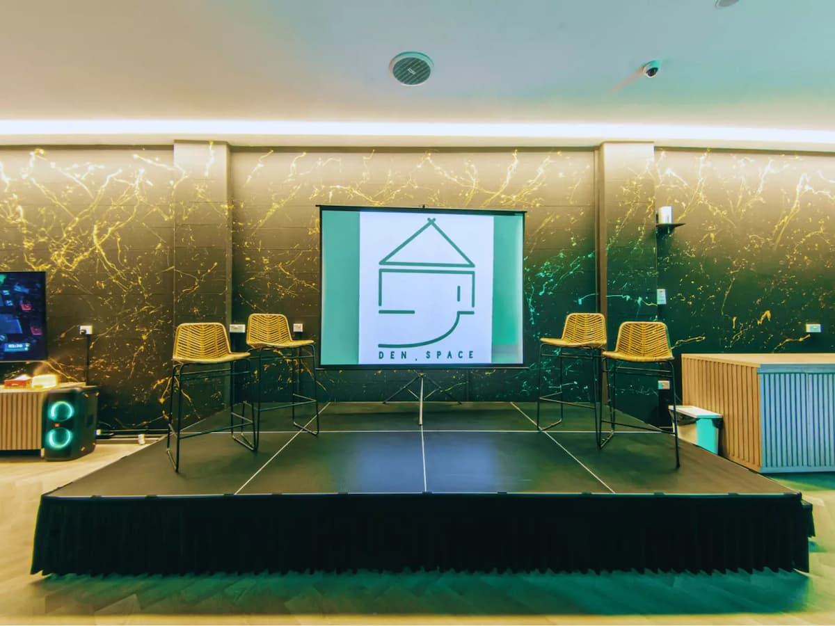 A small stage with two wicker chairs and a large screen displaying a minimalist logo reading "DEN SPACE.” The room has modern, elegant decor with marble-patterned walls.