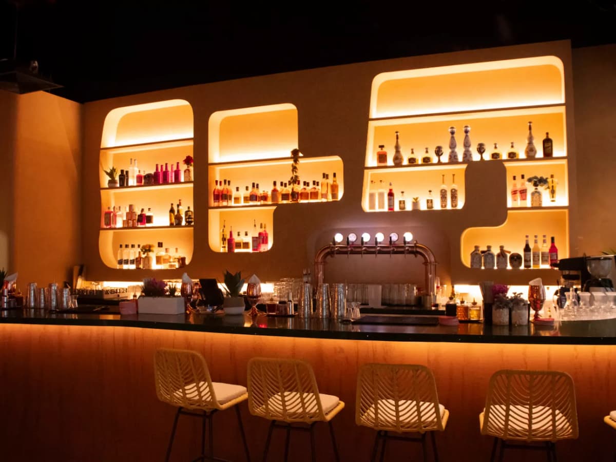 A stylish, warmly lit bar with modern shelves displaying vibrant bottles and cozy rattan bar stools