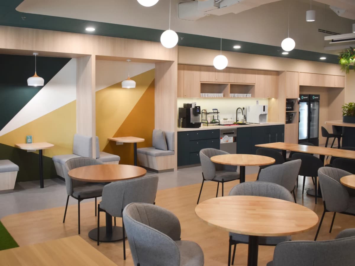 A modern coworking café-style lounge featuring round wooden tables, cushioned seating booths with geometric wall accents, and a fully equipped pantry area at the back.
