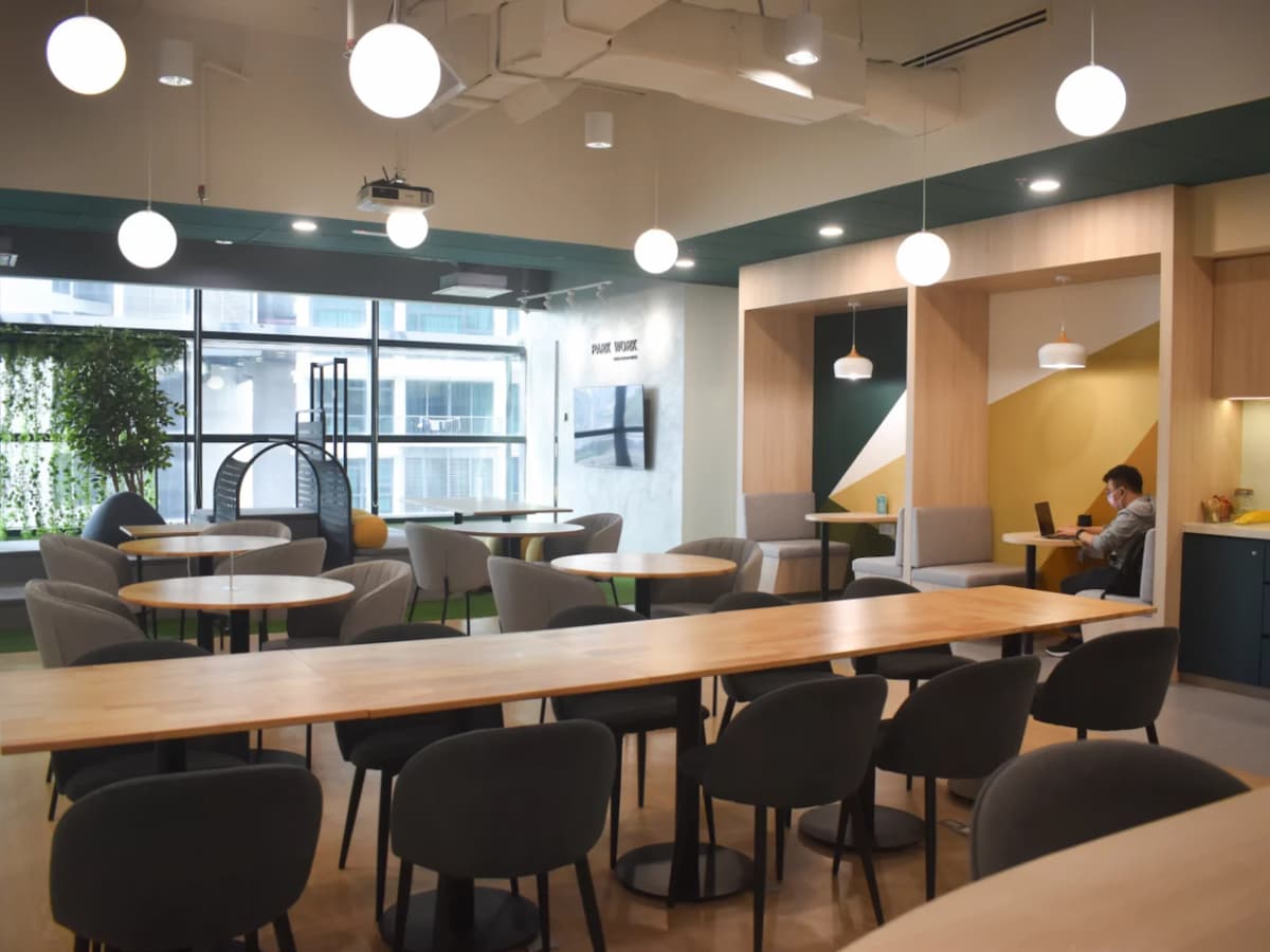 A spacious coworking lounge with long communal tables, round café-style seating, booth work pods, and large windows bringing in natural light, designed for casual collaboration and focused work.