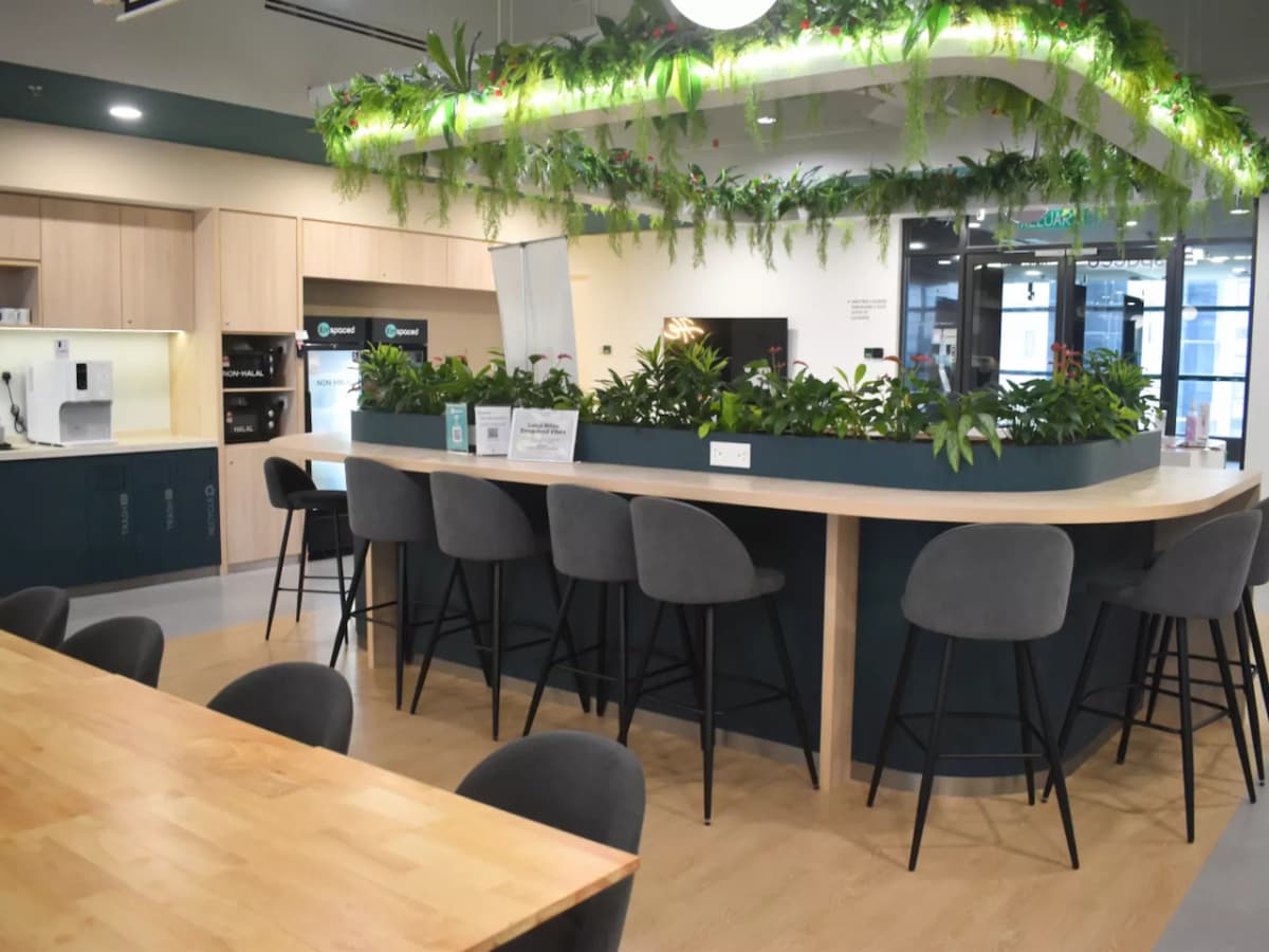 A modern coworking pantry area with a curved bar counter, high stools, lush greenery integrated into the design, and built-in coffee machines and refrigerators in the background.