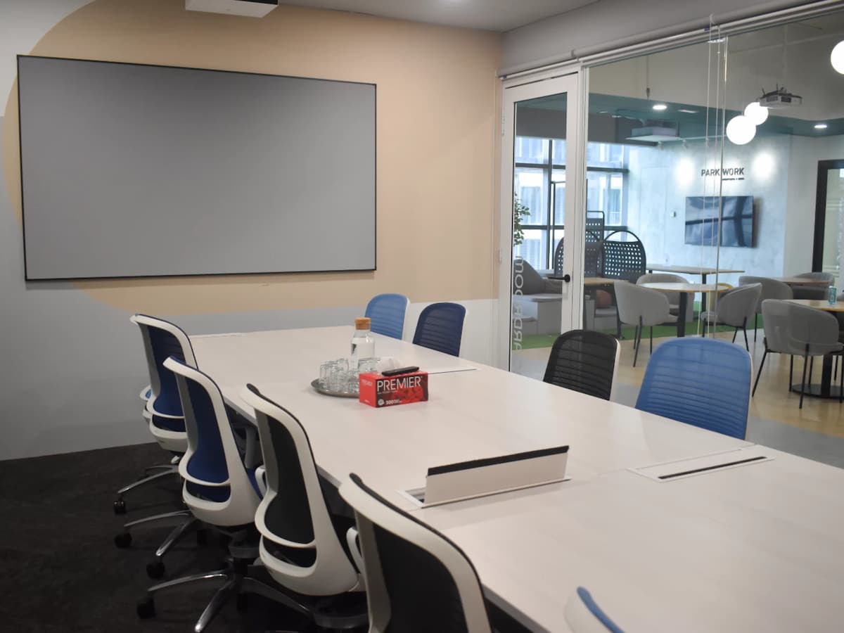 A sleek meeting room with a long white conference table, blue ergonomic chairs, a large wall-mounted screen, and glass doors opening to a coworking lounge area.