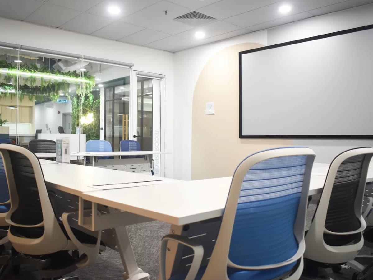 A contemporary meeting room with white tables, blue ergonomic chairs, a large wall-mounted screen, and glass walls overlooking a greenery-accented coworking space.