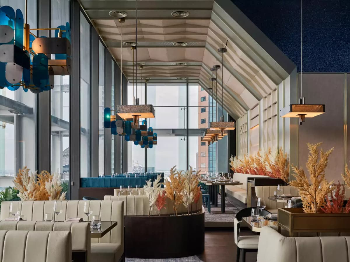 Sophisticated restaurant interior with curved cream booths, pendant lighting, decorative dried florals, and floor-to-ceiling windows overlooking the city.