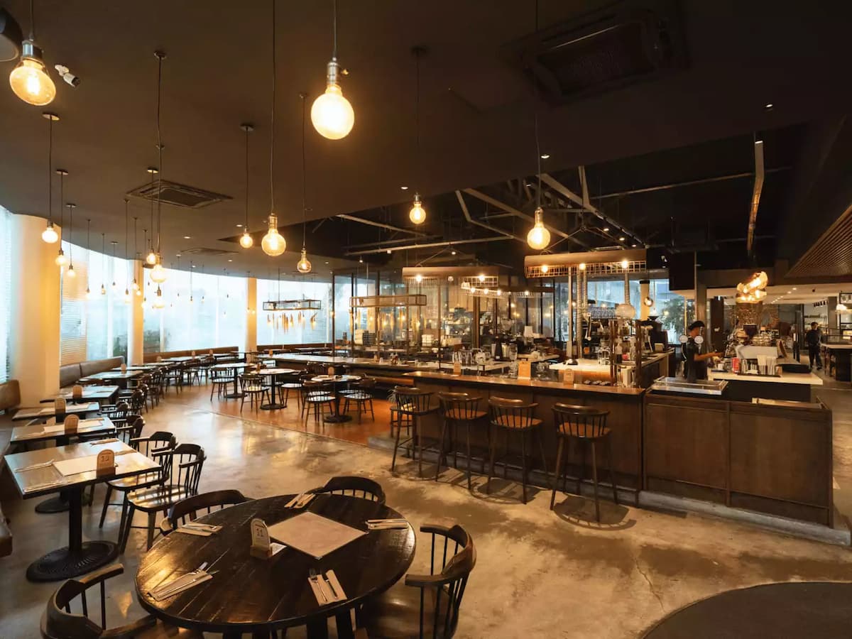 Industrial-style restaurant with central open kitchen, wooden bar seating, hanging Edison bulbs, and spacious dining area by large windows.