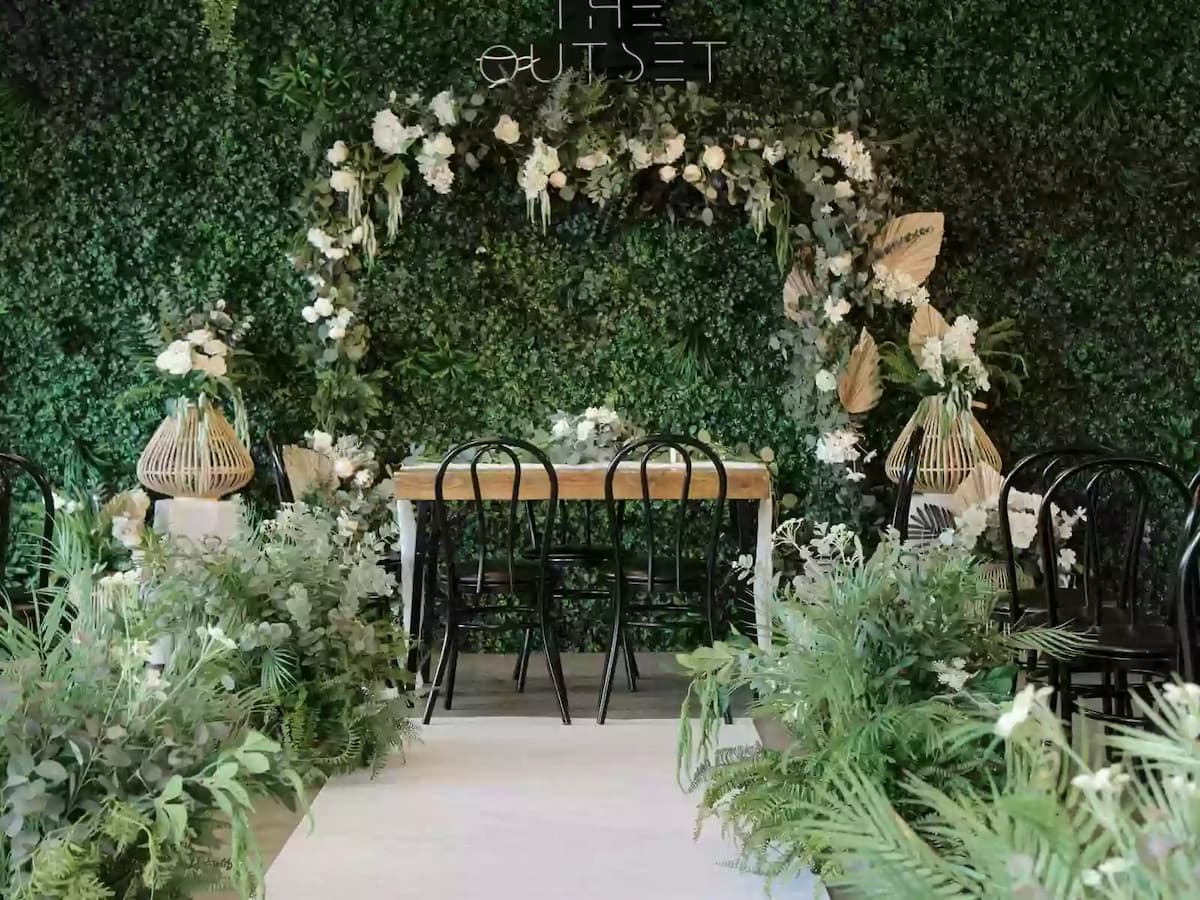 The Outset venue styled for a botanical wedding or solemnisation with a lush green living wall backdrop, floral arch, sweetheart dining table, black bistro chairs, rattan lanterns, and tropical greenery arrangements