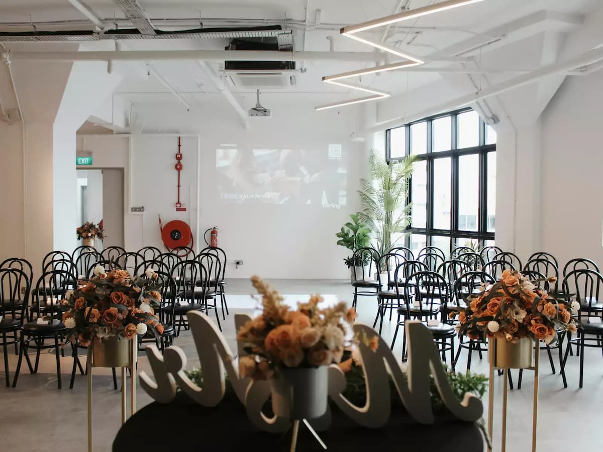 Wedding solemnisation setup in a bright white event space with rows of black bentwood chairs, terracotta floral arrangements on stands, a projection screen, and large industrial windows