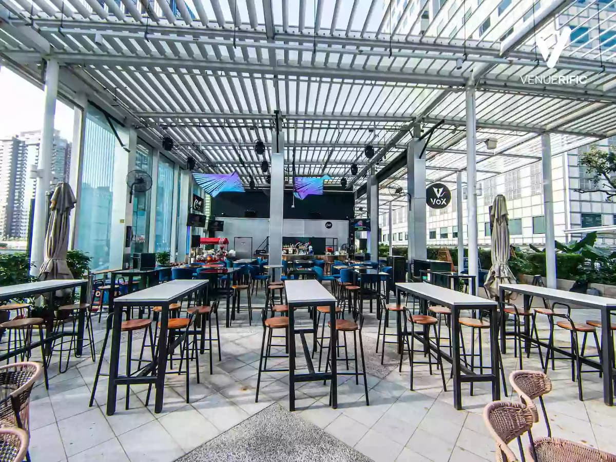 Spacious rooftop event space with high tables, blue chairs, bar area, and covered pergola with city views.