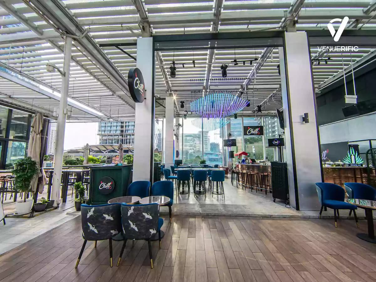 Open-air rooftop bar with blue velvet chairs, wooden flooring, LED ceiling lights, and a city skyline view.