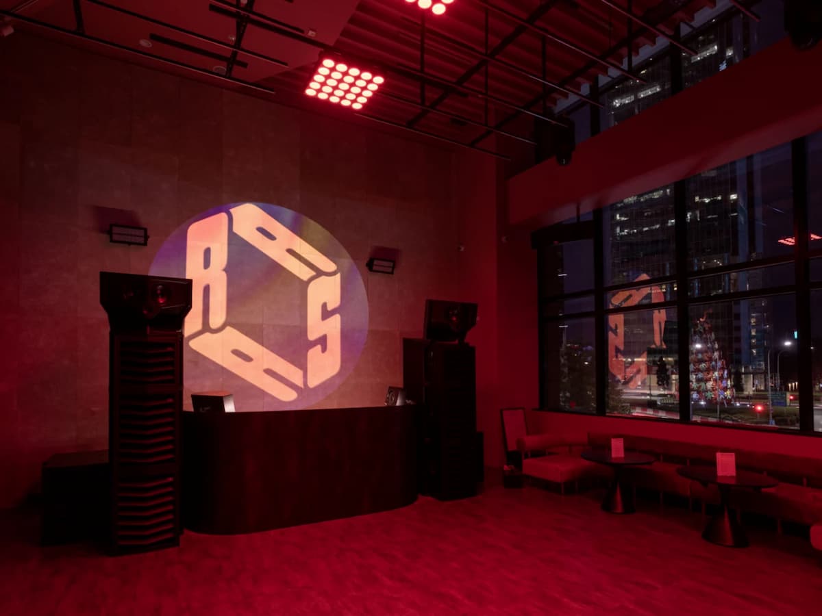 DJ booth with large speakers, red lighting, and city skyline view through floor-to-ceiling windows.