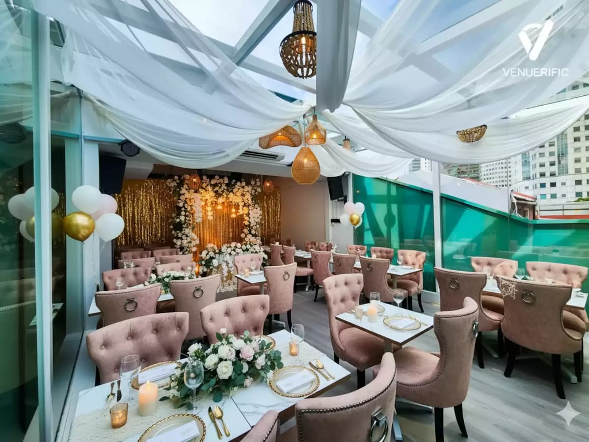 Elegant rooftop event space with blush pink chairs, white draped ceiling, floral backdrop, and gold-accented table settings.
