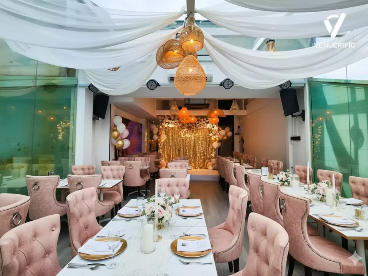 Elegant rooftop event space with blush pink chairs, white draped ceiling, floral backdrop, and gold-accented table settings.