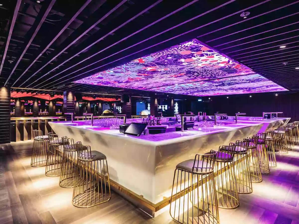 Modern bar with a glowing central counter, LED ceiling display, and sleek high stools in a vibrant nightlife setting.