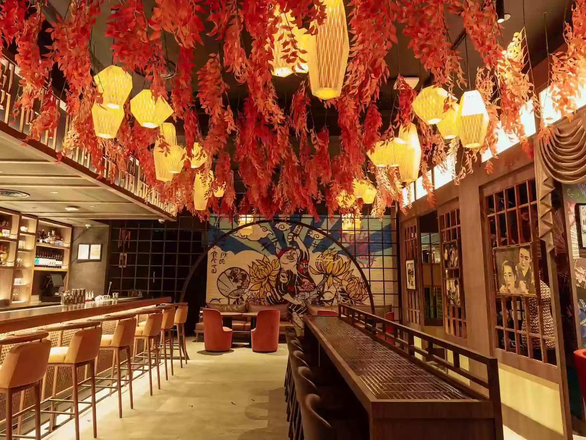 Atmospheric bar interior with hanging red foliage, warm lantern lighting, mural artwork, and bar seating.