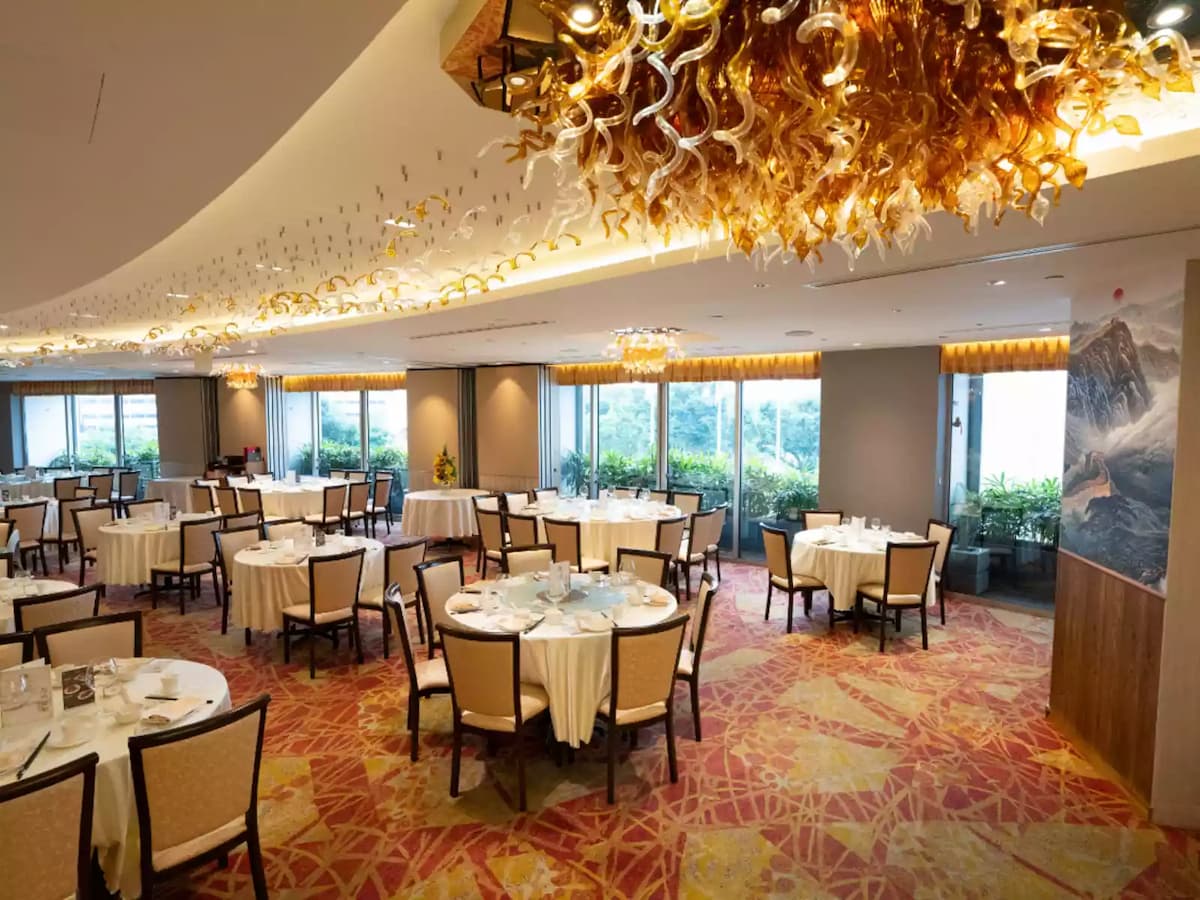 Elegant banquet hall with round dining tables, warm chandeliers, patterned carpet, and large windows overlooking greenery.