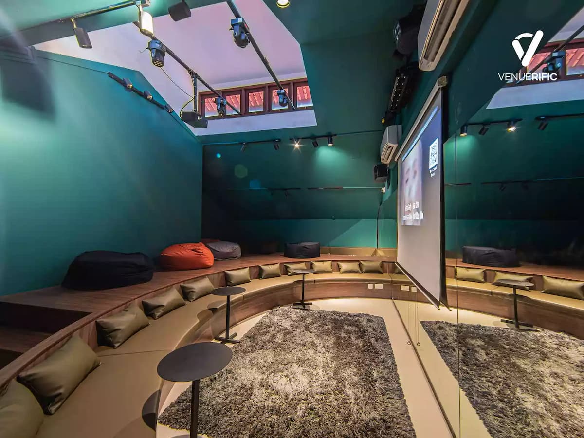 Intimate karaoke lounge with curved bench seating, scatter cushions, bean bags, a projection screen, shag rug, and teal walls with stage lighting above