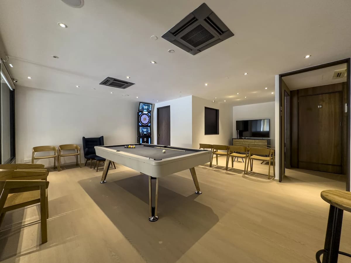 A minimalist recreational room featuring a light-colored pool table, a digital dartboard, and wooden seating against clean white walls.