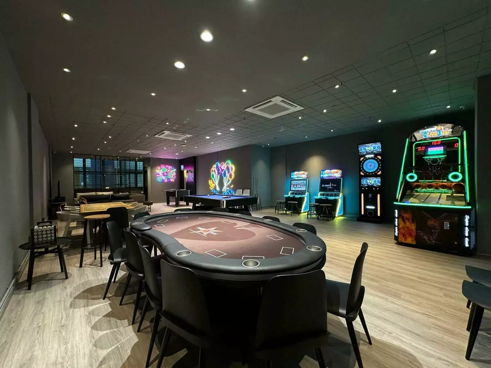 A vibrant entertainment room featuring a central poker table, pool table, and various arcade games under cool-toned lighting.