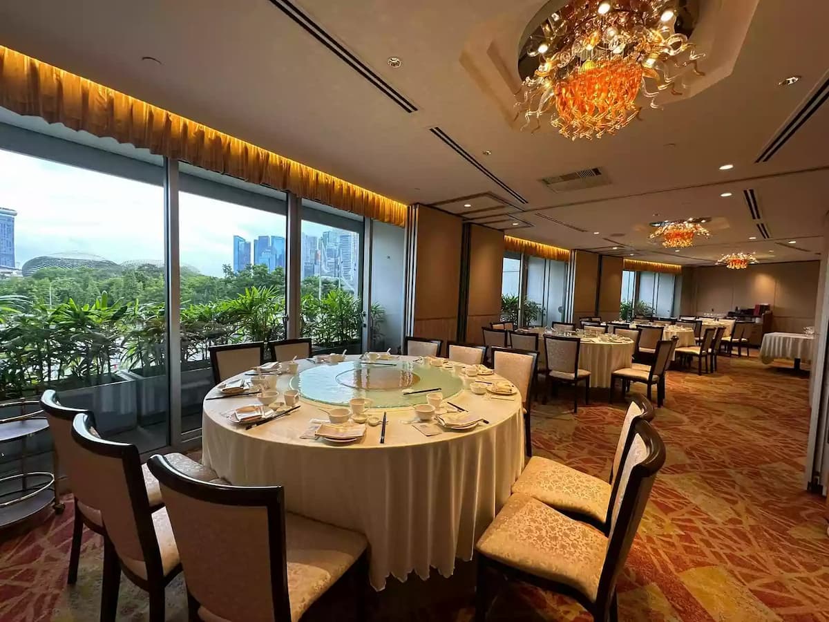 Bright banquet dining space with round tables, elegant chandeliers, patterned carpet, and panoramic city views through large windows.
