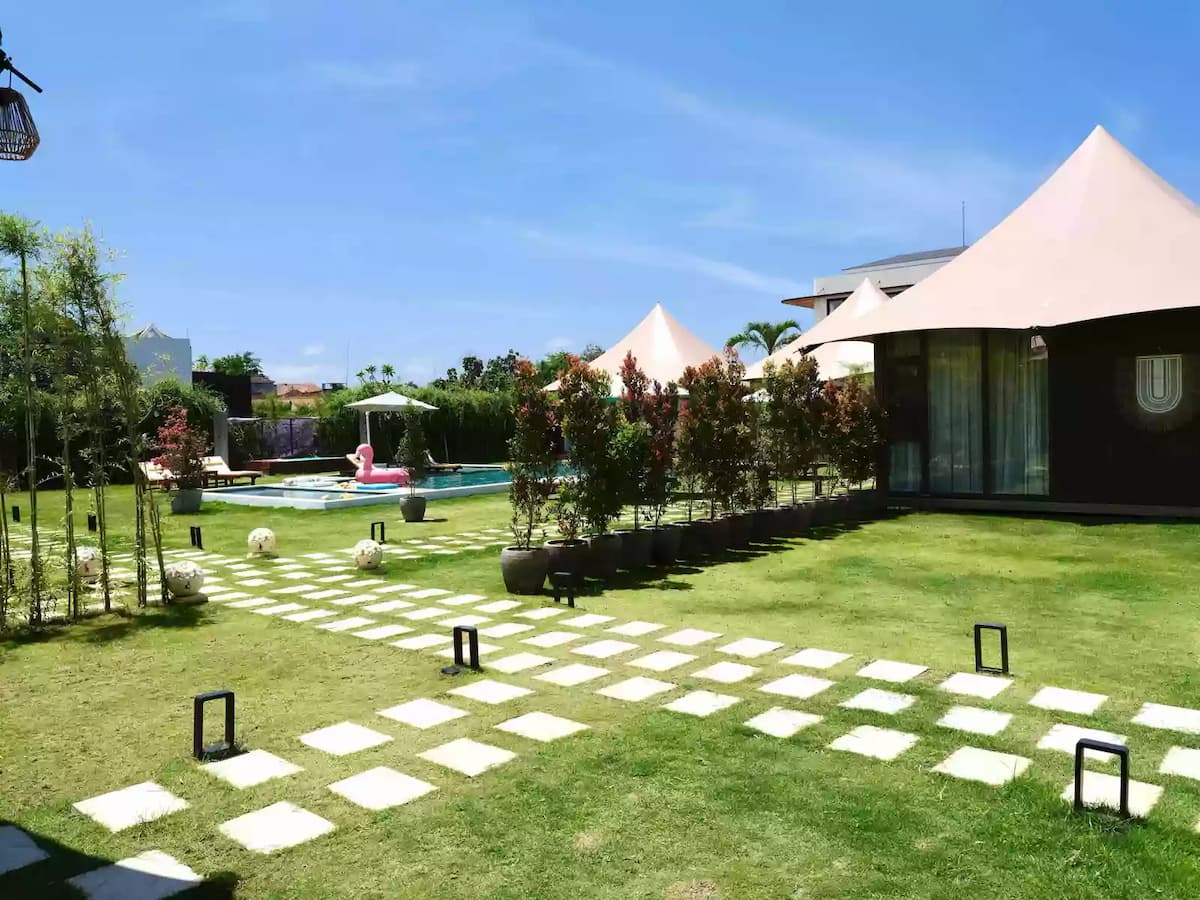 Resort-style garden with lawn pathways, pool area, and modern tented villas under a clear blue sky.