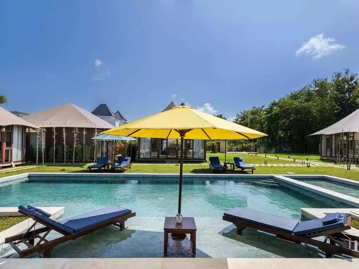 Poolside loungers beneath a yellow umbrella with modern villas and landscaped lawn under a clear blue sky.