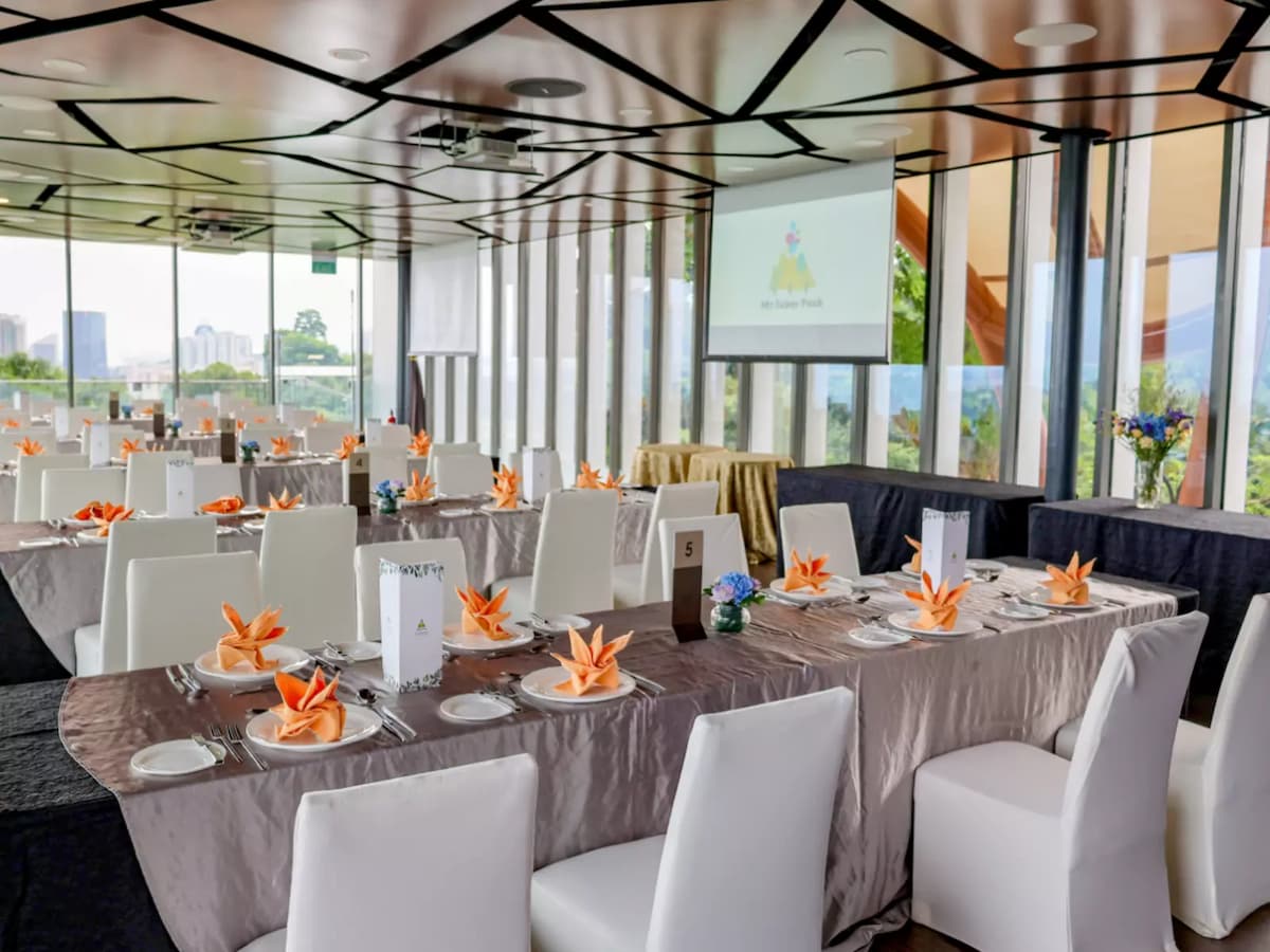 Bright event dining room with long banquet tables, white chairs, orange folded napkins, and a presentation screen by floor-to-ceiling windows.