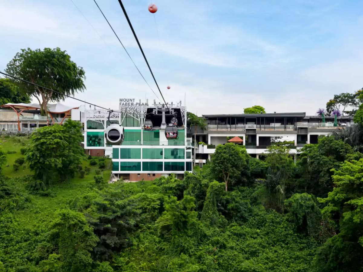 Cable car cabins arriving at Mount Faber Peak station above lush green hillside under a clear blue sky.