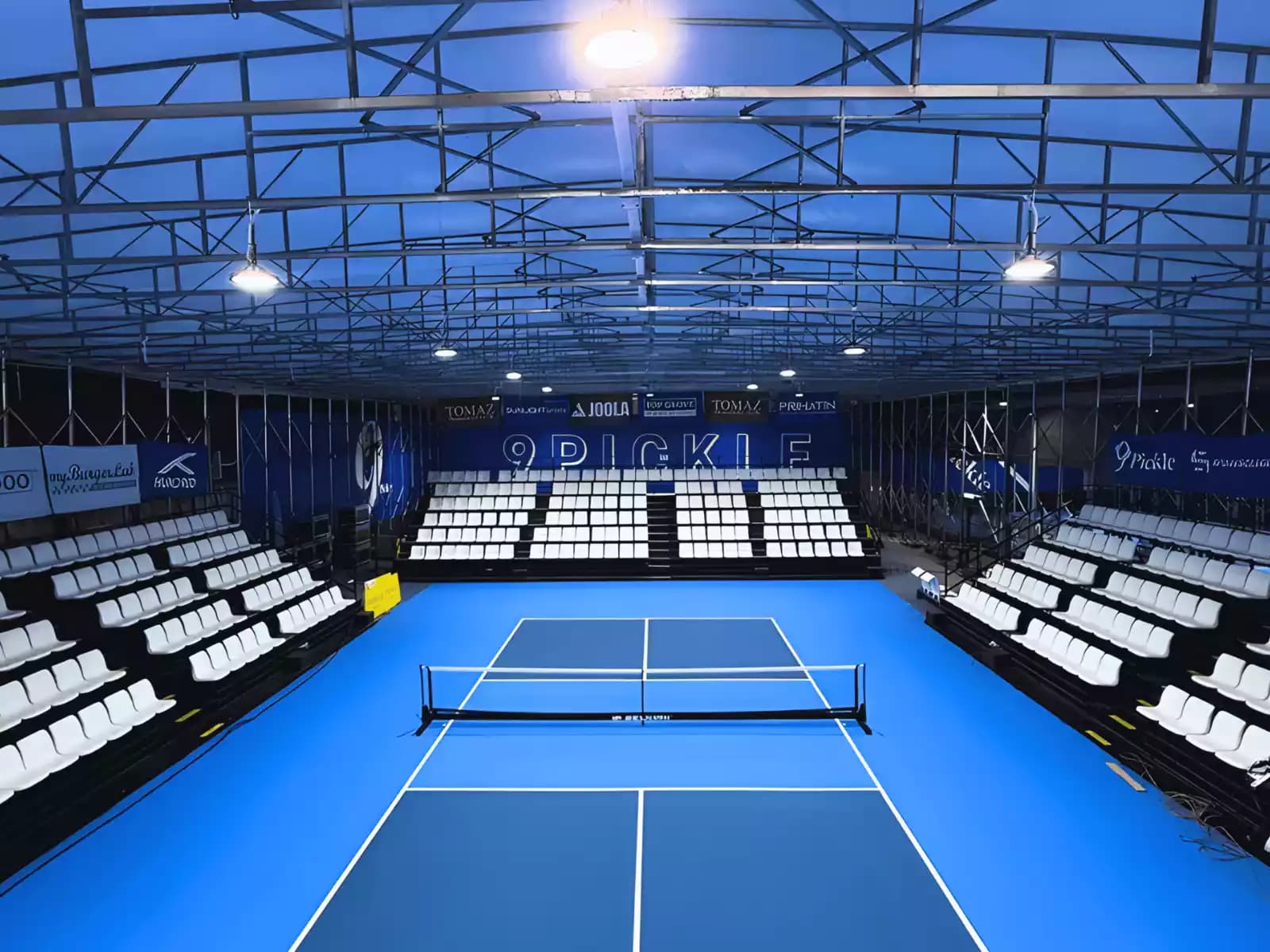 Indoor pickleball stadium with blue courts, spectator seating on both sides, and overhead lighting under a metal roof.