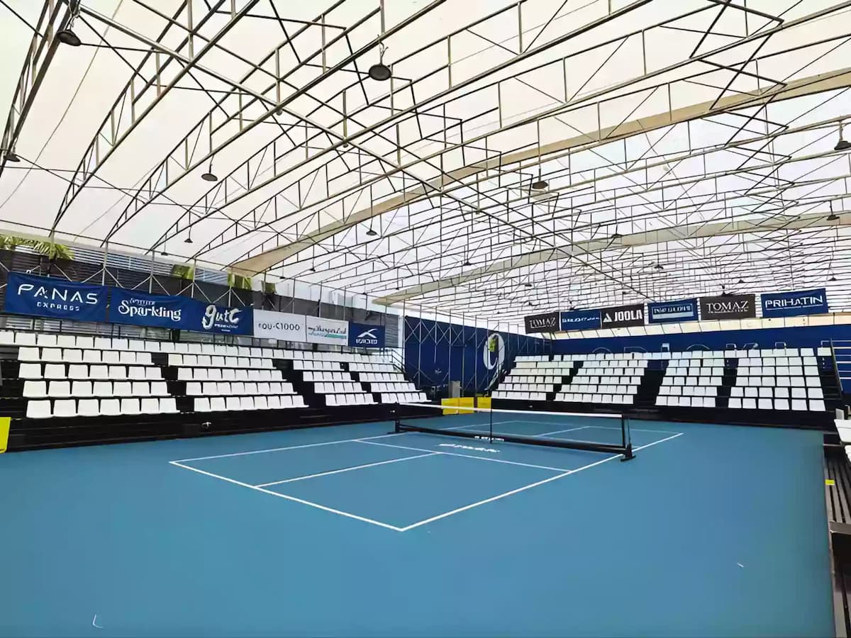 Indoor pickleball court with blue playing surface, spectator seating, and covered roof structure for tournaments and matches.