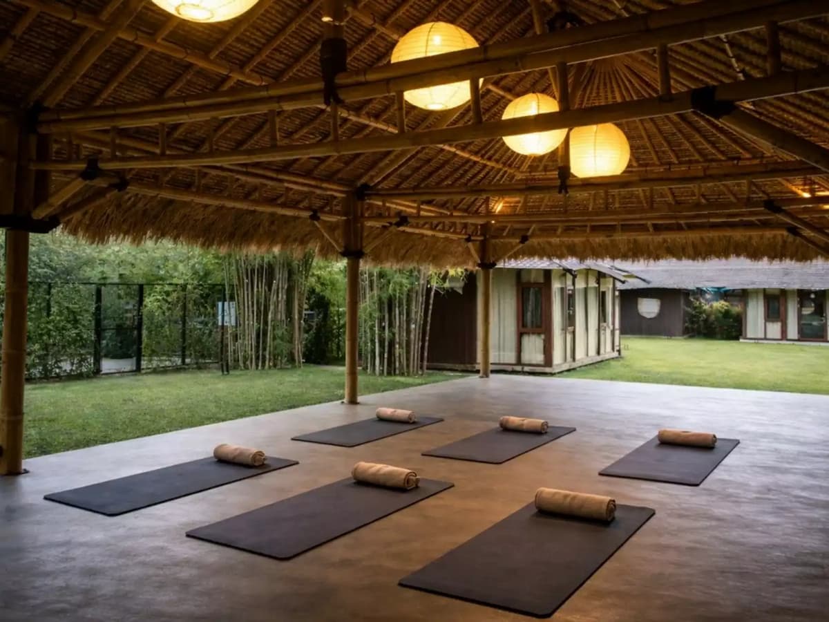 Open-air yoga pavilion with thatched roof, hanging lanterns, and mats arranged on a wooden deck overlooking a garden.