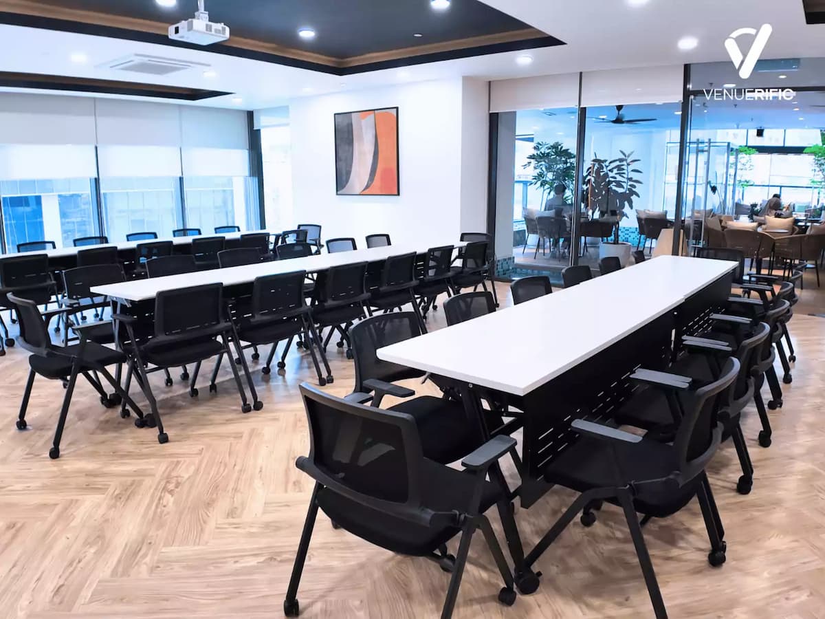 Modern meeting room with long white tables, black office chairs, and glass walls overlooking a lounge area.